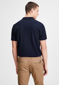 Marineblaues Polo-Shirt mit kurzen Ärmeln, aus weichem Baumwollstoff. Der Träger ist von hinten zu sehen und trägt khakifarbene Hosen mit zwei Gesäßtaschen.