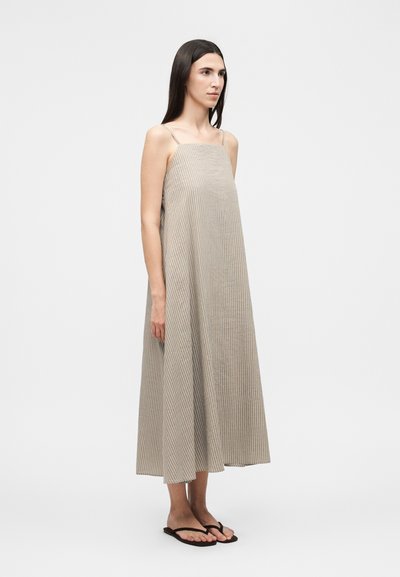 Γυναίκα που στέκεται πλαγίως, φοράει αμάνικο, φαρδύ φόρεμα μήκους έως τον αστράγαλο σε μπεζ χρώμα με λεπτές ρίγες και μαύρα σανδάλια, σε απλό φόντο.