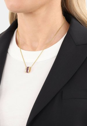 Collana d'oro con un pendente che presenta due bande intrecciate, una delle quali è decorata con pietre viola, indossata sopra una camicia bianca e un blazer nero.