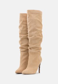 Bottes en daim beige montantes avec un design loose, un bout pointu et un talon aiguille présentant un accent texturé sur le talon.