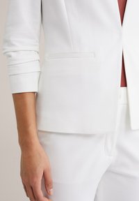 Valkoinen räätälöity bleiserikangas, jossa on sileä rakenne, yksittäinen etutasku ja käännetyt kalvosimet. Alla näkyy tummanpunainen paita.