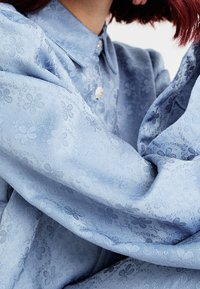 Světle modrá košile vyrobená z texturované tkaniny s květinovým vzorem. Ozdobena klasickým límcem a předním zapínáním na knoflíky.