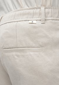 Calças de algodão bege com um bolso traseiro liso, cintura elástica e pequeno detalhe com etiqueta da marca. Tecido texturizado com padrões subtils.