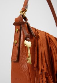 Brązowa skórzana torebka z zamszowymi frędzlami, złotymi okuciami i akcentem w postaci klucza. Posiada gładką teksturę oraz zorganizowany kształt.