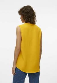 Top giallo senza maniche con un design testurizzato, orlo arrotondato e una leggera lucentezza, abbinato a pantaloni in denim blu con borchie decorative.