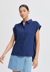 Camicia navy a maniche corte con colletto strutturato, tessuto texturizzato e spacchi laterali, indossata con una gonna in denim azzurro chiaro.