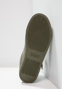 Suela de zapatilla verde oliva con surcos texturizados, que presenta una forma suave y redondeada y un logotipo estampado en el talón. Se observan bordes reforzados.