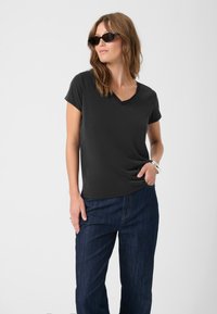 Černé tričko s výstřihem do V vyrobené z jemné tkaniny, s krátkým rukávem, nošené s tmavými denimovými kalhotami. Model pózuje s jednou rukou na boku a s brýlemi.