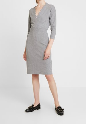 Kobieta ubrana w dopasowaną szarą dzianinową sukienkę z karbowanym dekoltem w kształcie litery V i rękawami do połowy przedramienia, w połączeniu z czarnymi mokasynami ozdobionymi złotymi klamrami.