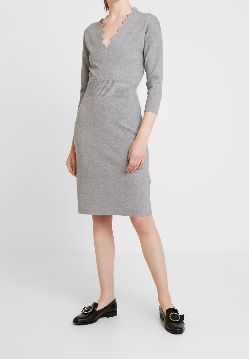 Femme portant une robe en maille grise ajustée avec un col en V festonné et des manches trois-quarts, associée à des mocassins noirs avec des boucles dorées.