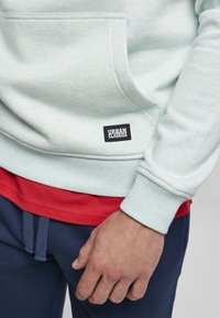 Felpa azzurro chiaro con tasca frontale, polsini a coste e patch logo nero. Indossata sopra una maglietta rossa e pantaloni blu scuro.