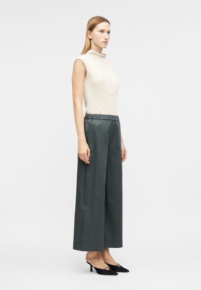 Γυναίκα που φοράει μπεζ τοπ χωρίς μανίκια με ψηλό λαιμό, φαρδιά σκούρα γκρι παντελόνια και μαύρες γόβες με μυτερή μύτη και χαμηλό τακούνι, σε προφίλ.