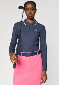 Marineblå piké med lange ermer og hvit krage, kombinert med et rosa skjørt og blått belte. Hånd som holder et golfklubbe, teksturert stoff.