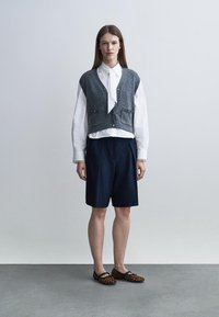 Femme debout portant un chemisier blanc avec cravate, un gilet sans manches gris, un short marine à hauteur des genoux et des ballerines imprimées léopard.