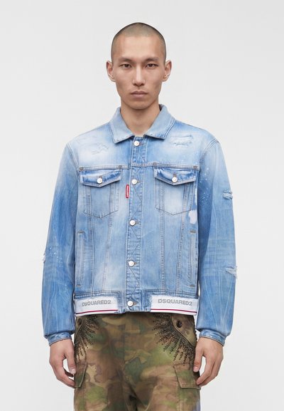 Homme portant une veste en jean bleu délavée avec des détails usés et un pantalon cargo camouflage, debout devant un fond gris clair.