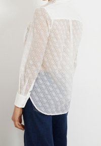 White sheer blouse with a diamond pattern, long sleeves, and a collar. Features button cuffs and a curved hem, paired with dark blue jeans.