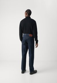 Svart langermet skjorte, blå denimjeans og svarte sko. Jeansene har et brunt lærlap på livlinen og en rett ben-design.