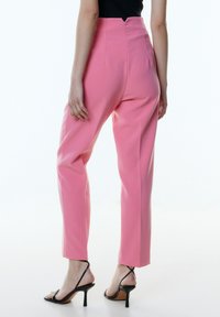 Roze high-waisted broek met een aangesloten pasvorm, met een gladde textuur en subtiele kreukdetails, gecombineerd met zwarte sandalen met banden.