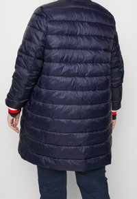 Tmavě modrý prošívaný péřový kabát, délka ke kolenům, dlouhé rukávy, s červenobílými pruhovanými akcenty na manžetách. Hladká textura.