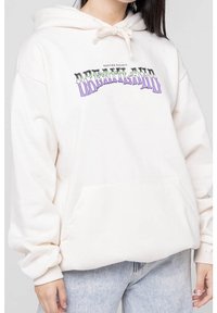 Bílá mikina s kapucí a šňůrkou, na přední straně s fialovým a černým grafickým textem "DREAMLAND" a přední kapsou. Měkká tkanina.