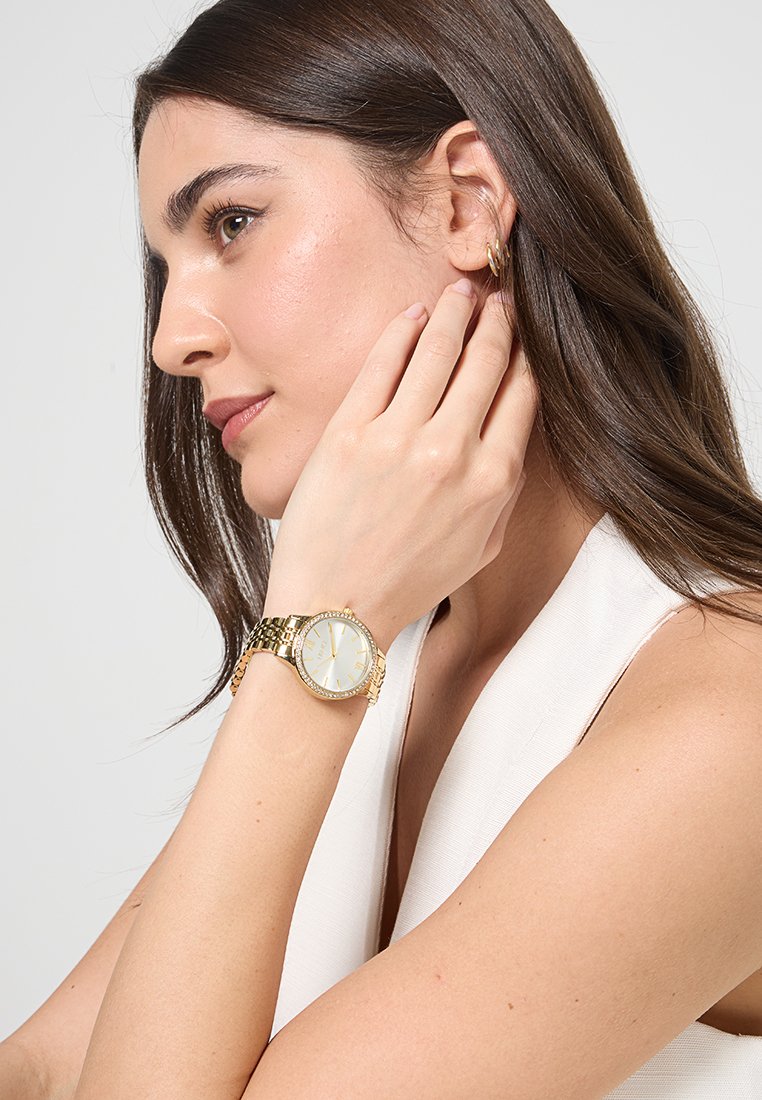 Orologio da donna in oro con quadrante bianco rotondo, cinturino testurizzato e lunetta gioiello, indossato su un polso. Design semplice ed elegante.