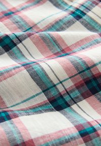 Tessuto di cotone intrecciato con un motivo a quadri composto da linee rosse, turchesi e blu scuro su uno sfondo crema, che mostra la texture e una leggera caduta.