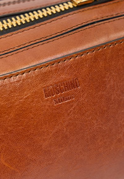 Καφέ δερμάτινη τσάντα με ανάγλυφο λογότυπο "MOSCHINO Milano" και χρυσή λεπτομέρεια φερμουάρ με μαύρο υφασμάτινο τελείωμα.