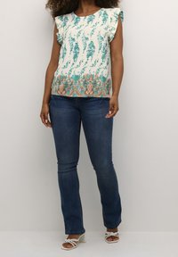 Femme portant un haut blanc à motifs floraux teal, un jean bleu et des sandales à talons blancs, se tenant contre un mur léger et uni.