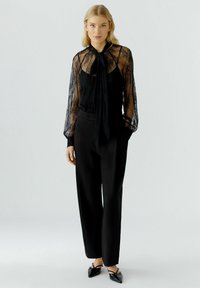 Schwarze Spitzenbluse mit hohem Kragen und transparenten langen Ärmeln, kombiniert mit hochgeschnittenen schwarzen Hosen und spitzen schwarzen Schuhen.