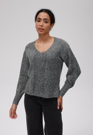 Maglione grigio a maglia con scollo a V, polsini a costine e un motivo testurizzato al centro. Indossato sopra pantaloni neri a gamba larga.