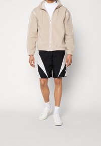 Person som bär en beige fleece zip-up hoodie, vit polotröja, svart och vit sportshorts, vita strumpor och vita kardborresandaler.