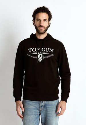 Mand med krøllet hår og skæg iført sort hættetrøje med tekst og emblem "TOP GUN", kombineret med lyseblå jeans, stående mod hvid baggrund.