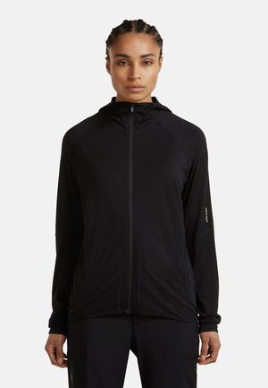 Vrouw met gevlochten haar draagt een zwart zip-up hooded jack en zwarte broek, staand tegen een effen witte achtergrond.