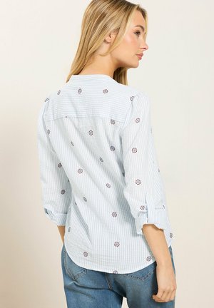 Vrouw in lichtblauw gestreept shirt met kleine bloemenprints, opgerolde mouwen, blauwe spijkerbroek, kijkt naar rechts tegen een effen achtergrond.