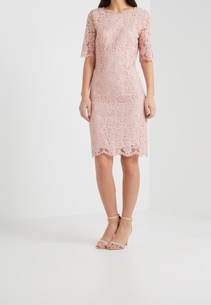 Femme portant une robe en dentelle rose mi-longue avec des manches coude et des talons nus à bride cheville, debout devant un fond uni.