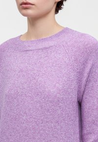 Gros plan d'une personne portant un pull tricoté violet doux avec un col rond côtelé et des manches raglan.