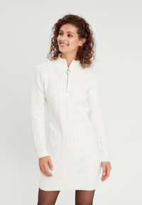 Robe tricotée blanche avec un col zippé, un design en tricot torsadé et des motifs en diamant. Manches longues avec des poignets et un ourlet côtelés.