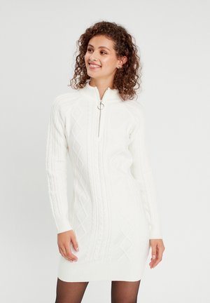 Robe tricotée blanche avec un col zippé, un design en tricot torsadé et des motifs en diamant. Manches longues avec des poignets et un ourlet côtelés.