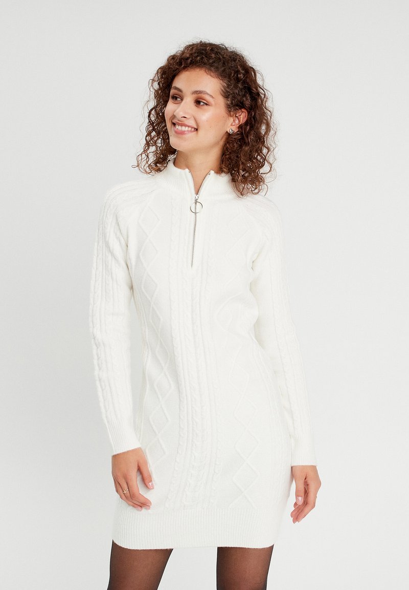 Robe tricotée blanche avec un col zippé, un design en tricot torsadé et des motifs en diamant. Manches longues avec des poignets et un ourlet côtelés.