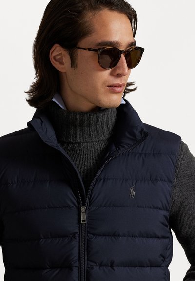 Navy puffer vest with a zip closure, worn over a dark gray ribbed turtleneck. Detailing includes a small logo on the chest and sunglasses.