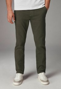Pantalones chinos verde aceituna de corte slim, confeccionados en mezcla de algodón, con bolsillos laterales y combinados con zapatillas blancas. Textura suave.