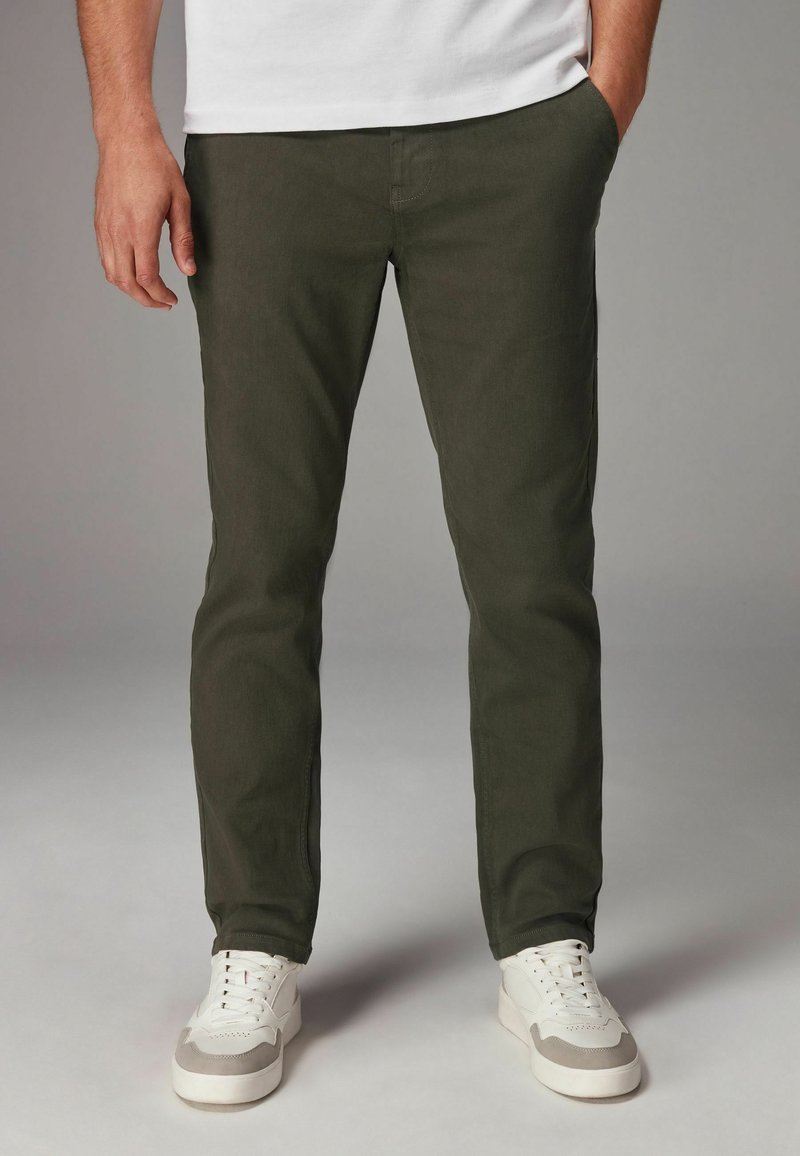 Pantalones chinos verde aceituna de corte slim, confeccionados en mezcla de algodón, con bolsillos laterales y combinados con zapatillas blancas. Textura suave.