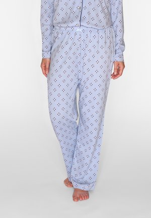 Personne portant un pantalon de pyjama bleu clair avec de petits motifs losanges foncés, pieds nus, debout devant un fond uni.