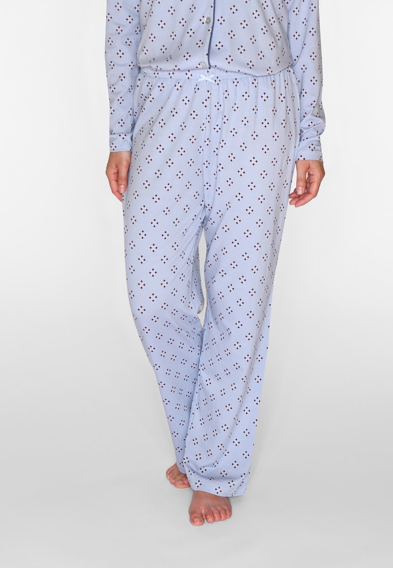 Personne portant un pantalon de pyjama bleu clair avec de petits motifs losanges foncés, pieds nus, debout devant un fond uni.