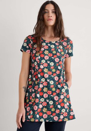 Vrouw in een tuniek met korte mouwen, voorzien van rode, roze, witte en olijfgroene bloemmotieven, staande tegen een effen lichte achtergrond.