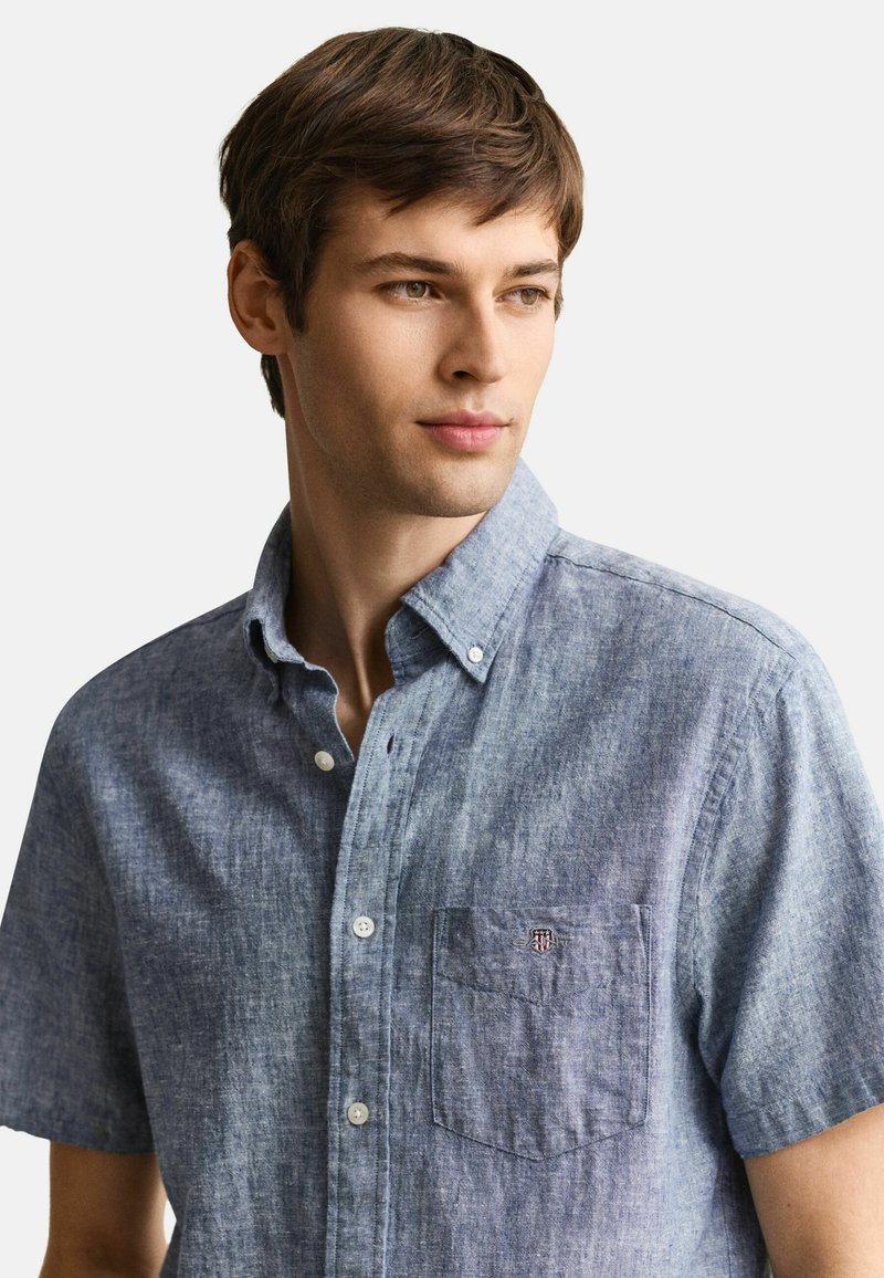 Giovane uomo con capelli castani che indossa una camicia blu a maniche corte con bottoni, che guarda leggermente verso destra su uno sfondo semplice.