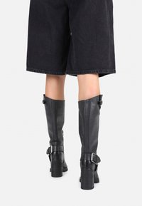 Stivali in pelle nera al ginocchio, con una texture liscia, tacchi imponenti e dettagli con fibbia. Mostrati indossati con pantaloni corti scuri e larghi.