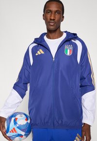 Modrá športová bunda s bielymi rukávmi a zlatými ozdobami, so znakom Talianska a dizajnom troch pruhov. Drží vzorovanú futbalovú loptu.