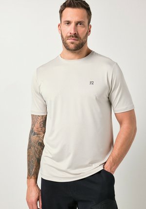 Mann mit Bart und detailliertem Tattoo am linken Arm, trägt ein hellbeiges Kurzarmhemd und dunkle Hose, steht mit der rechten Hand in der Hosentasche.