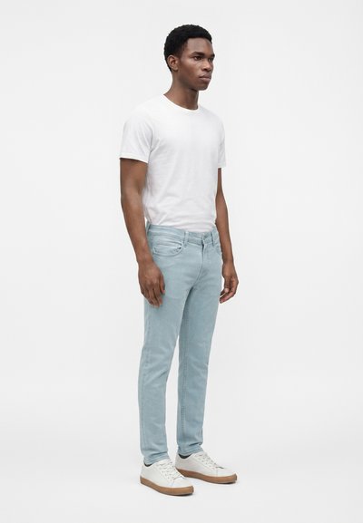 Homme aux cheveux courts et bouclés portant un t-shirt blanc, un jean bleu clair et des baskets blanches, debout devant un fond clair uni.